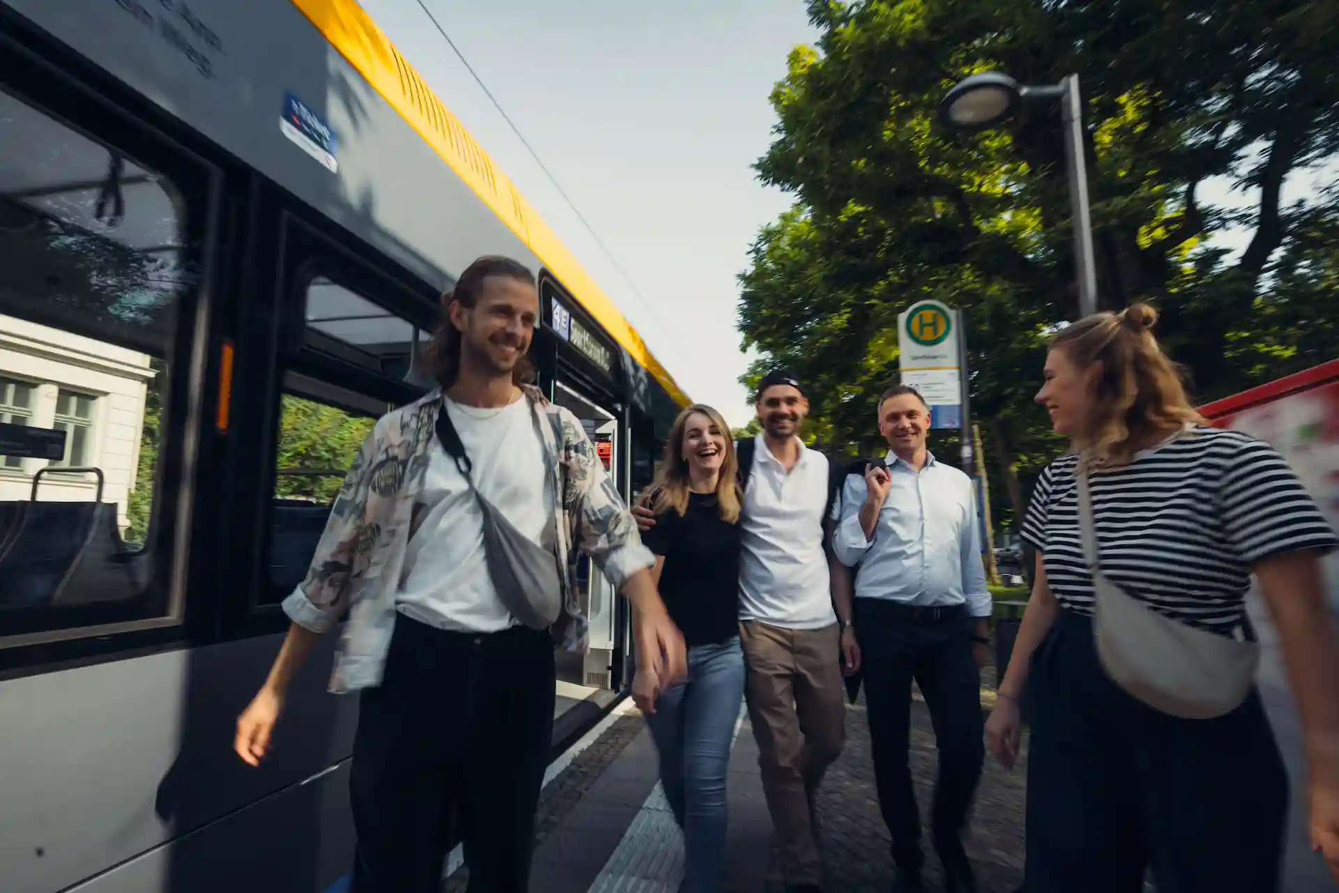 Fünf junge Erwachsene steigen lächelnd an einer sonnigen Bushaltestelle in der Stadt aus einem modernen Linienbus aus